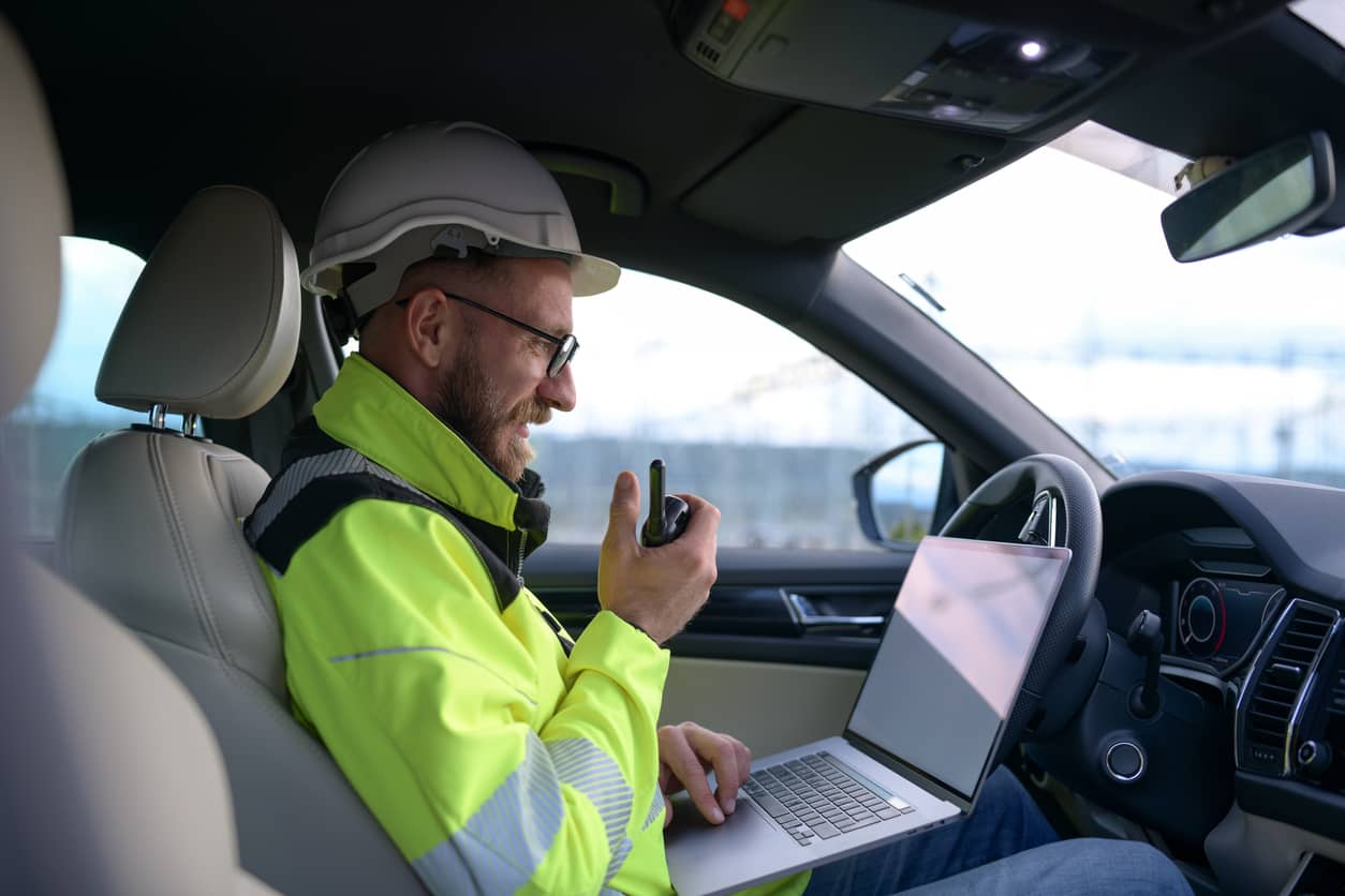 Using field service automation - Male engineer in his car working on different tasks on his laptop and holding a walkie-talkie.